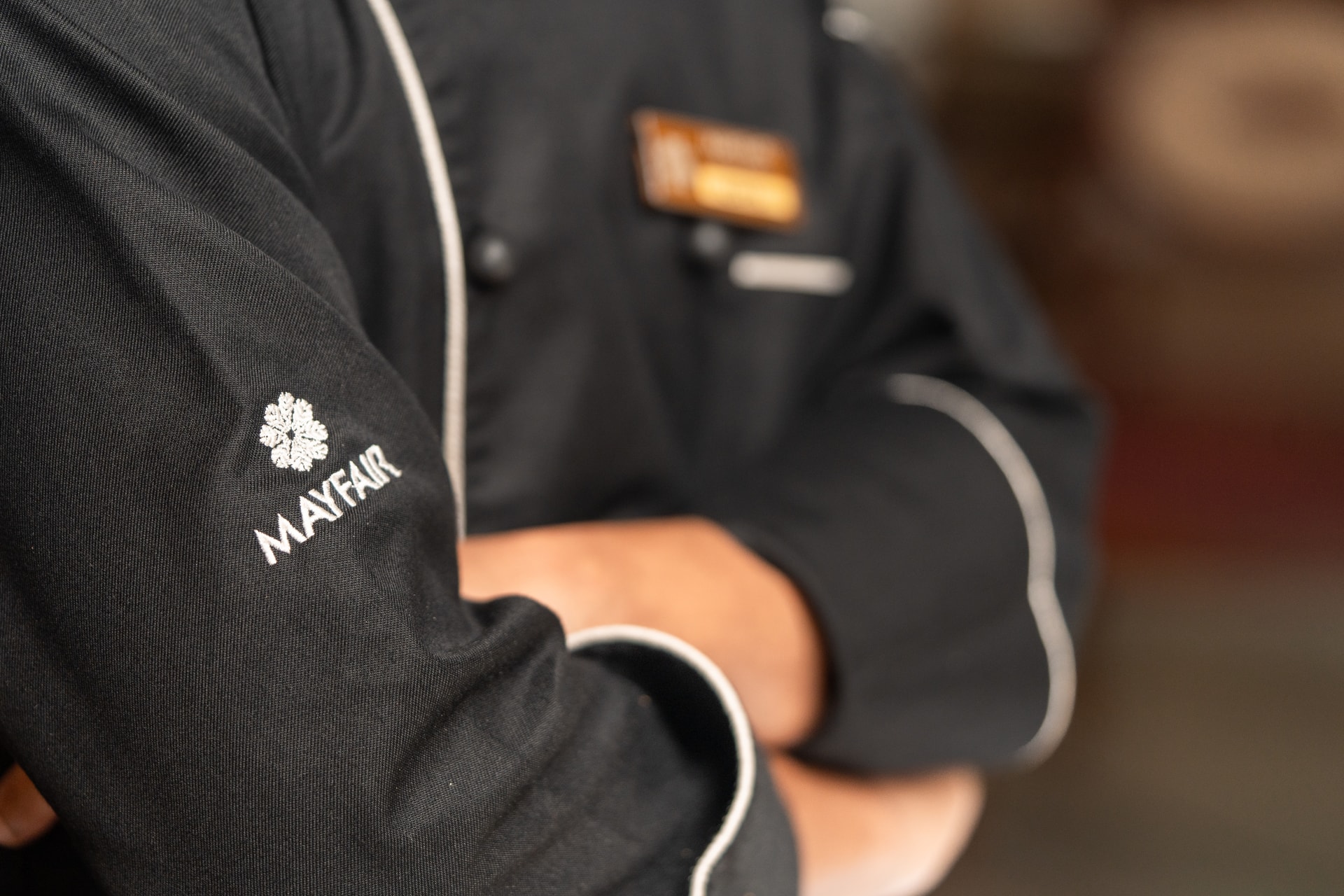 The MAYFAIR mark embroidered on the sleeve of a chef's black jacket, arms folded