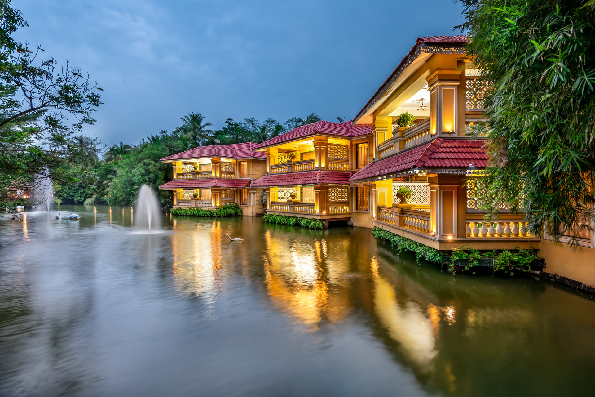 Luxury resort facade of MAYFAIR Lagoon Bhubaneswar illuminated at dusk reflecting on the lagoon with a fountain and swan