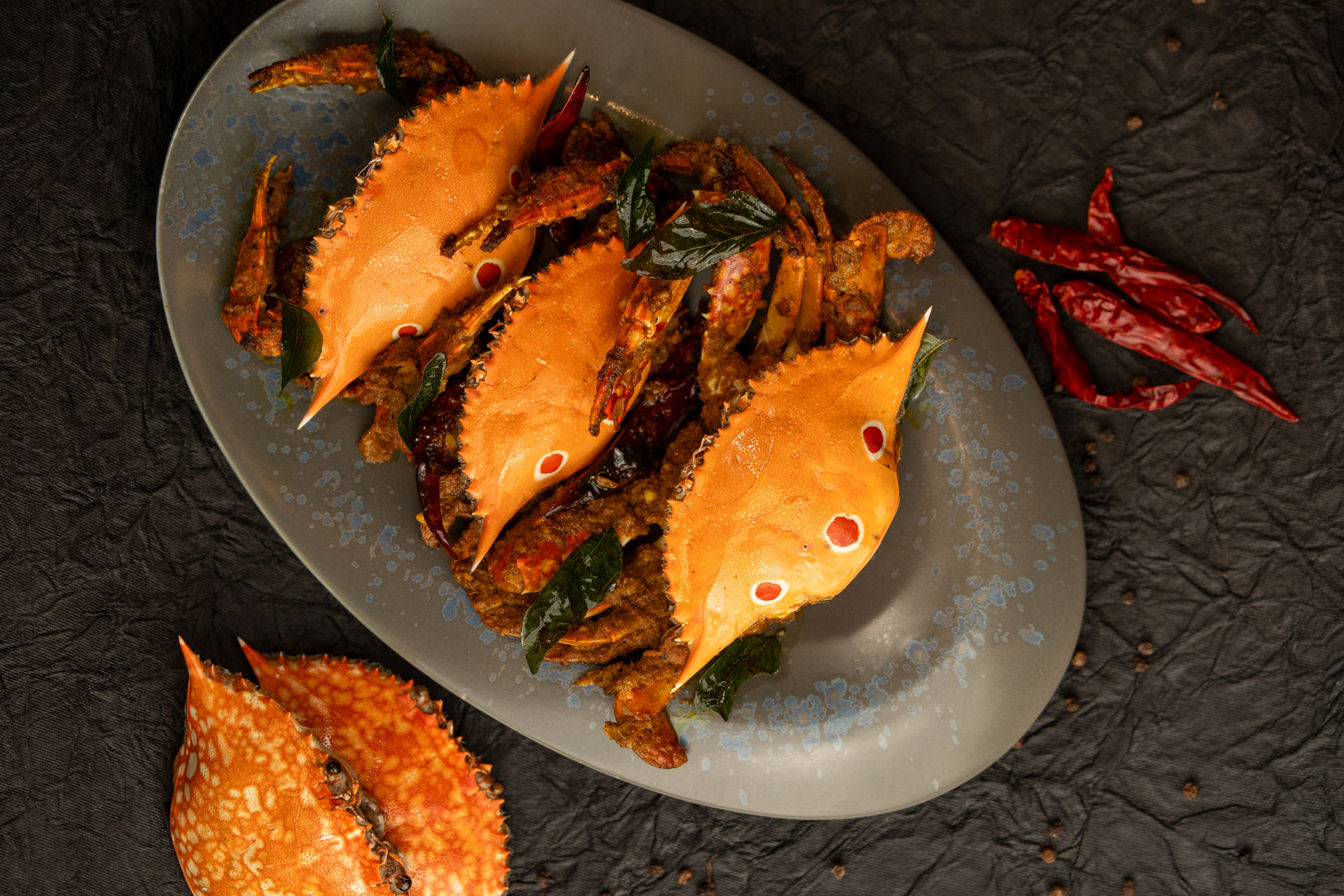 Crab in a dark spiced masala with curry leaves, three orange shells laid over it on a grey oval platter, dried red chillies alongside