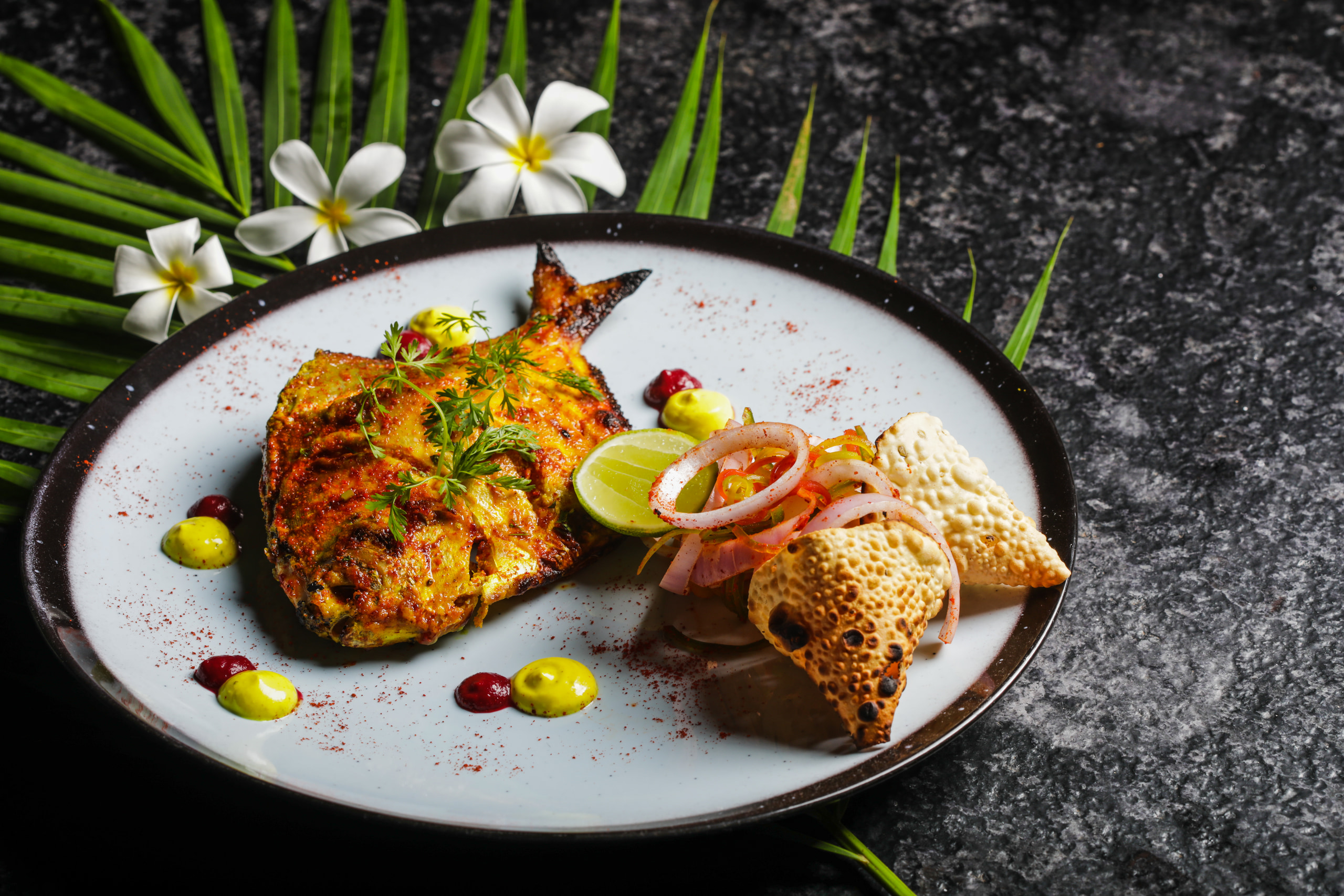 A whole tandoori fish plated with lime, onion and papad, on slate with frangipani