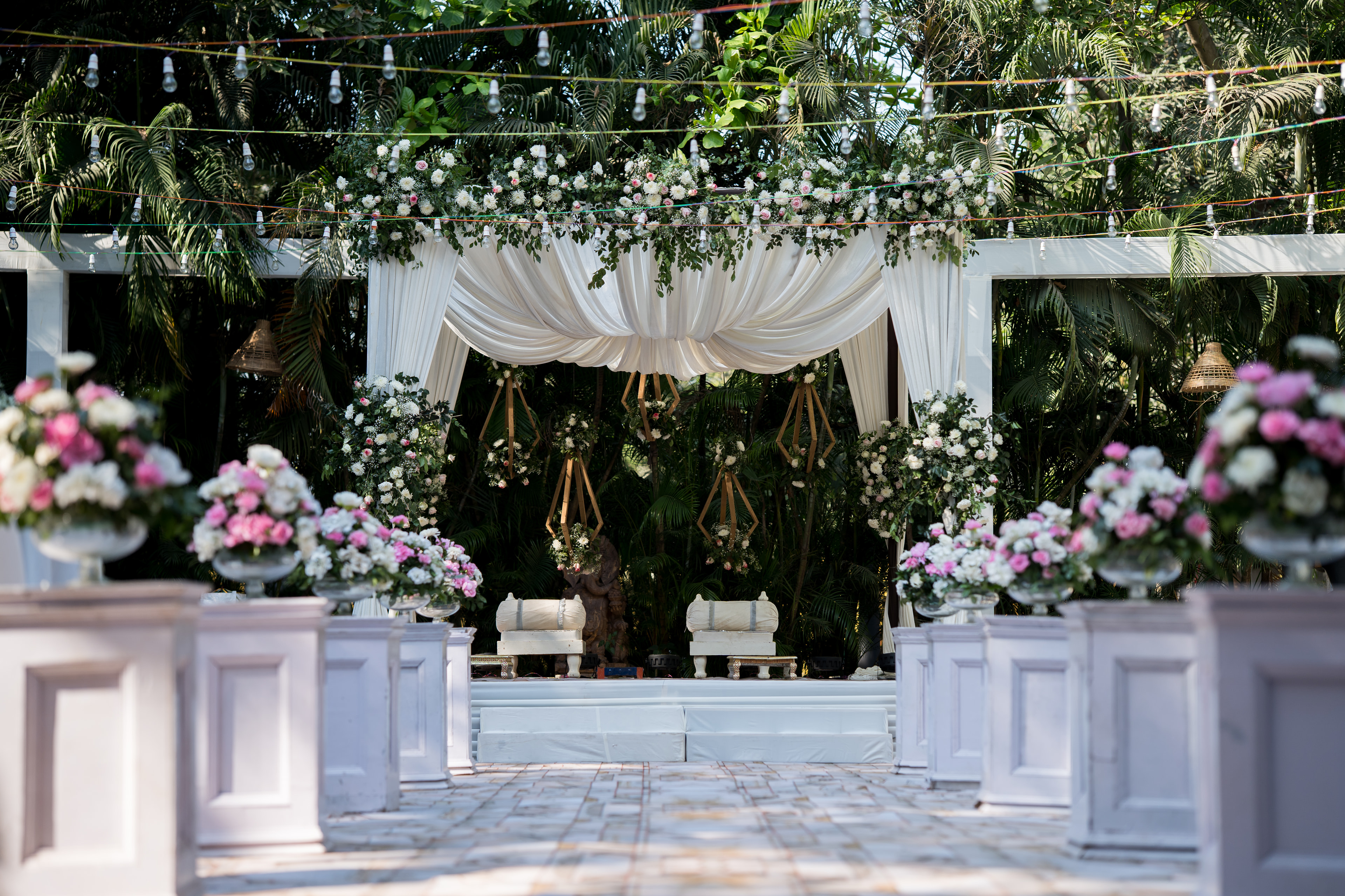 A reception stage dressed in white drapes, with pink and white flowers and strings of lights overhead