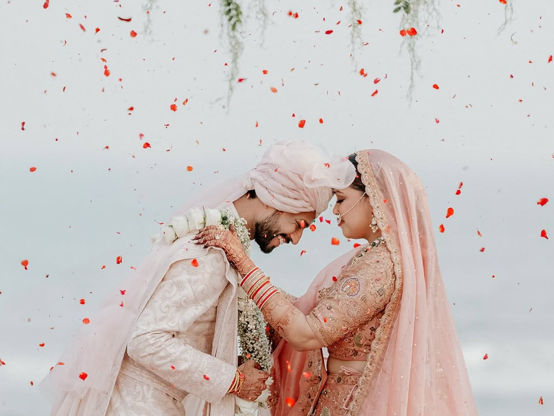 A bride and groom in pale pink, foreheads together under falling rose petals, the sea behind them
